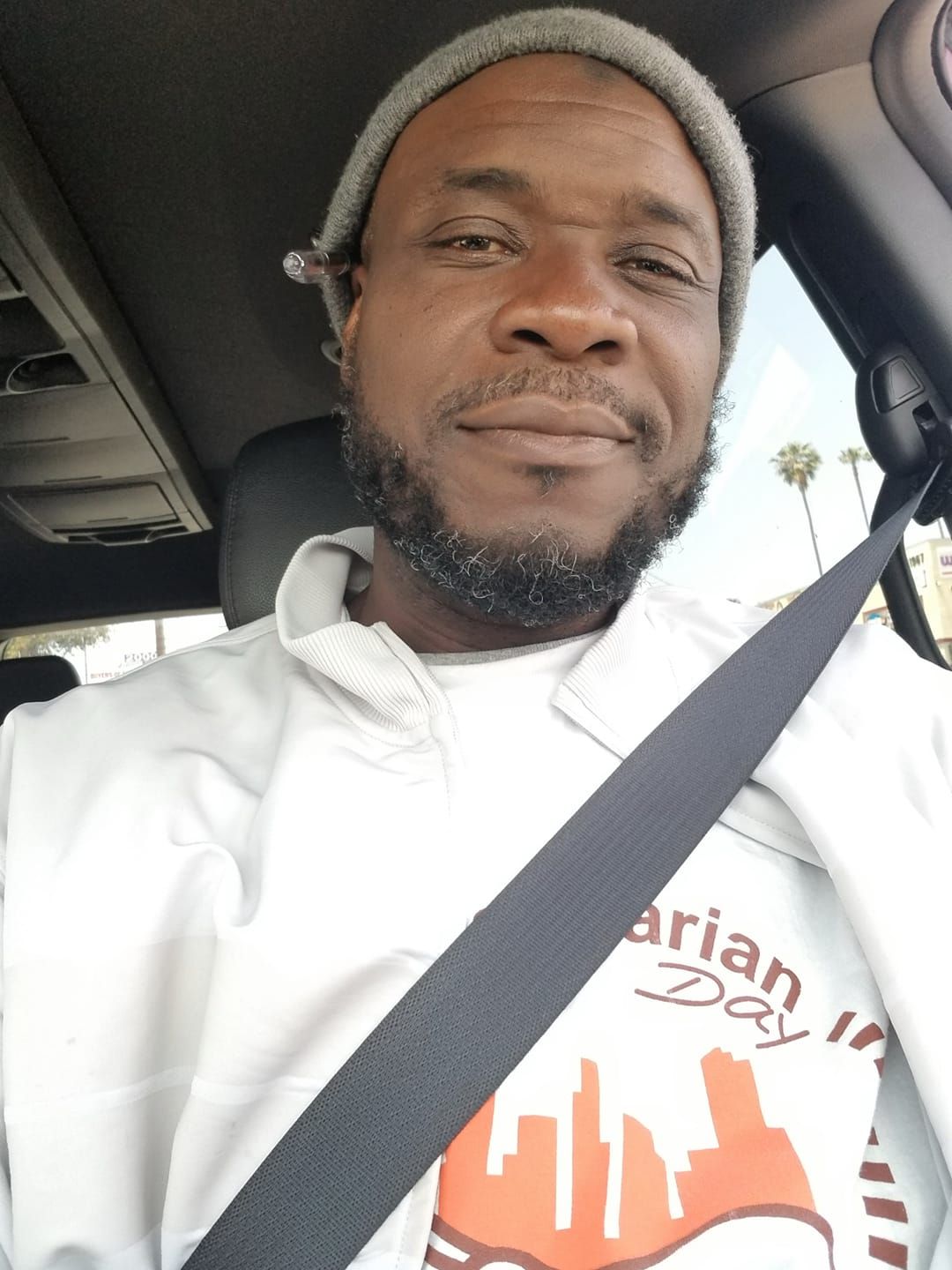 Man in a car, wearing a light-colored sweatshirt and beanie. He has a beard and is looking at the camera.