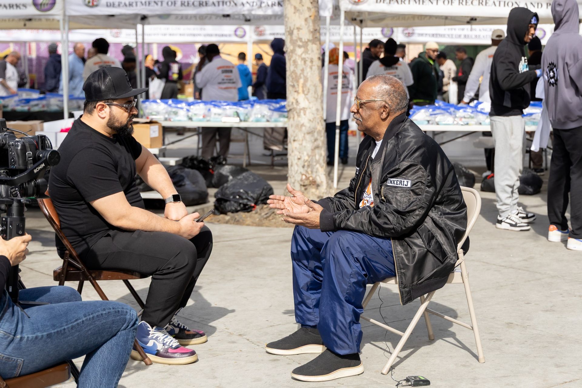 Two men seated, conversing outdoors at an event with tents and people in the background.