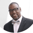 A Black man in a suit and bow tie, wearing glasses, smiling, possibly in a professional setting.