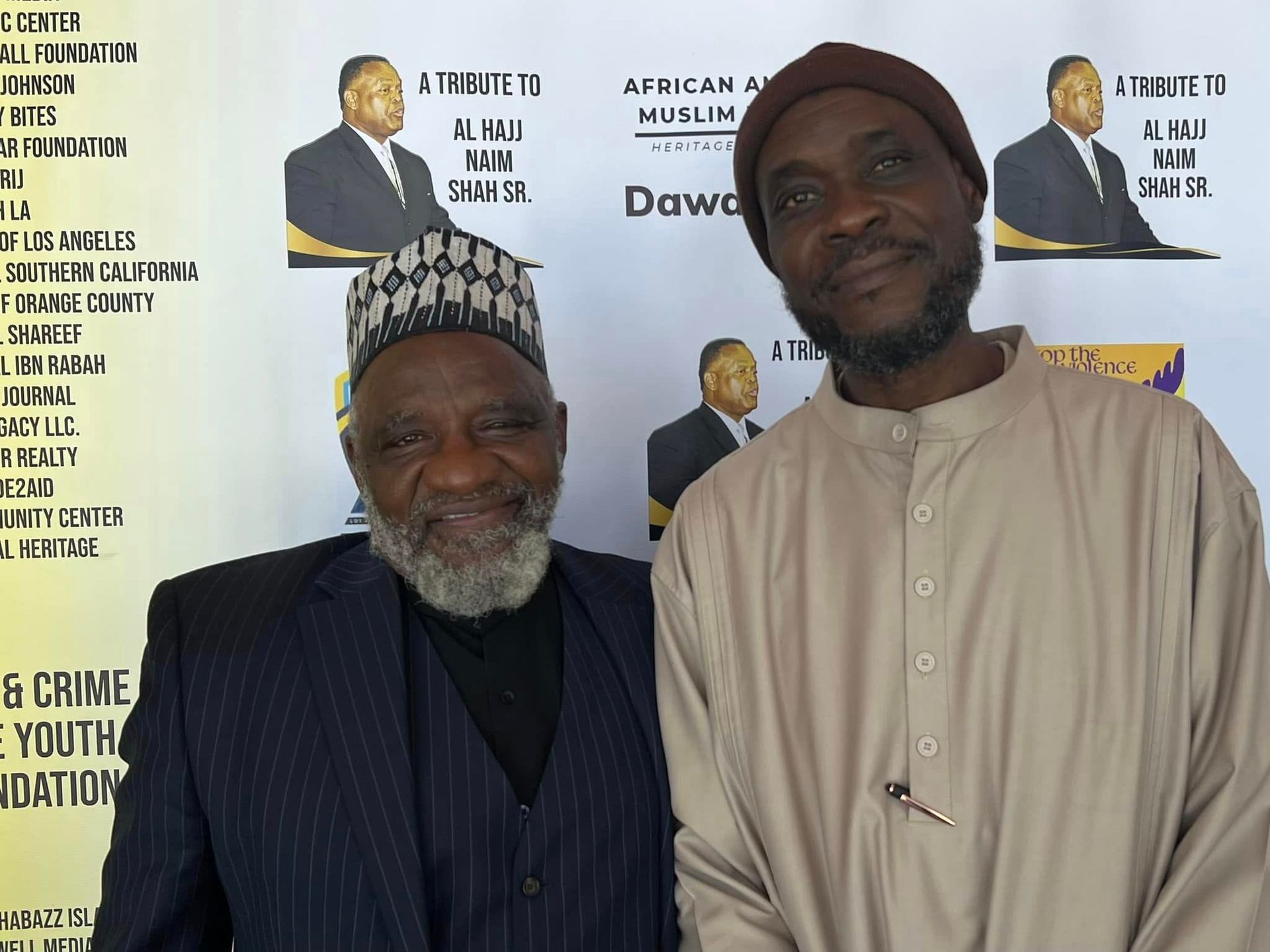 Two men pose together in front of banners. One wears a black suit, a patterned cap, and has a white beard. The other wears a light brown tunic and a beanie.