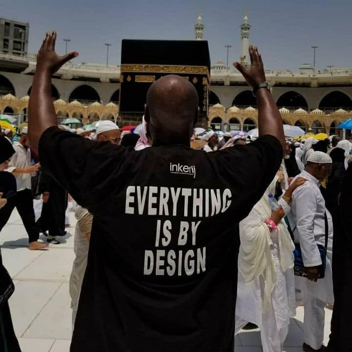 Man in black shirt with raised arms, in front of Kaaba. Shirt reads,