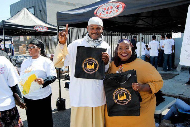People holding bags outdoors at an event; man raising a finger, woman smiling, tents in background.