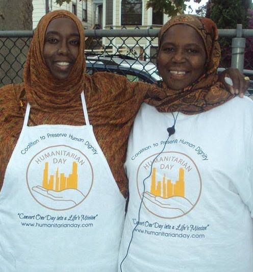 Two women in headscarves and Humanitarian Day shirts, smiling, outdoors. One wears an apron.