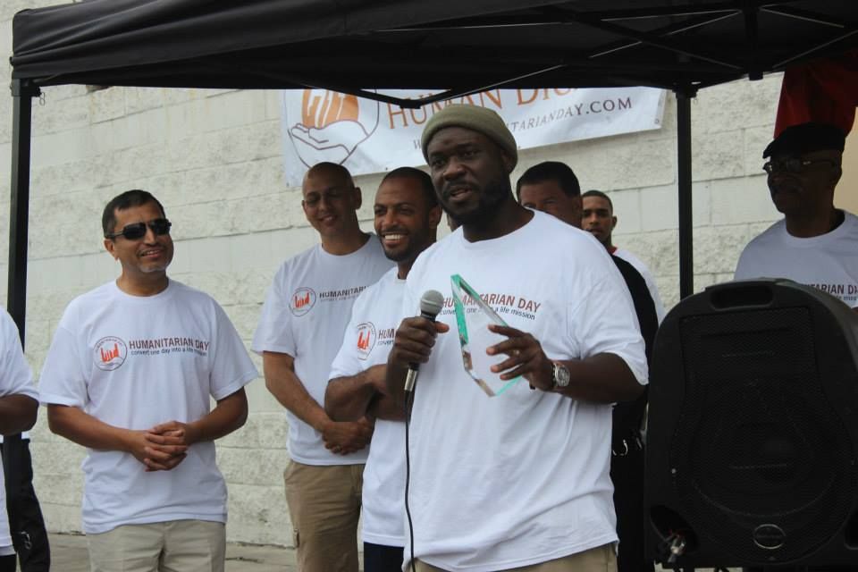 Man speaking into microphone, other men in white shirts stand behind him, outdoor event.
