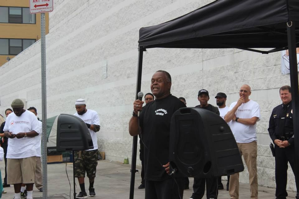 Man speaking into microphone, other people listen in front of a white building.