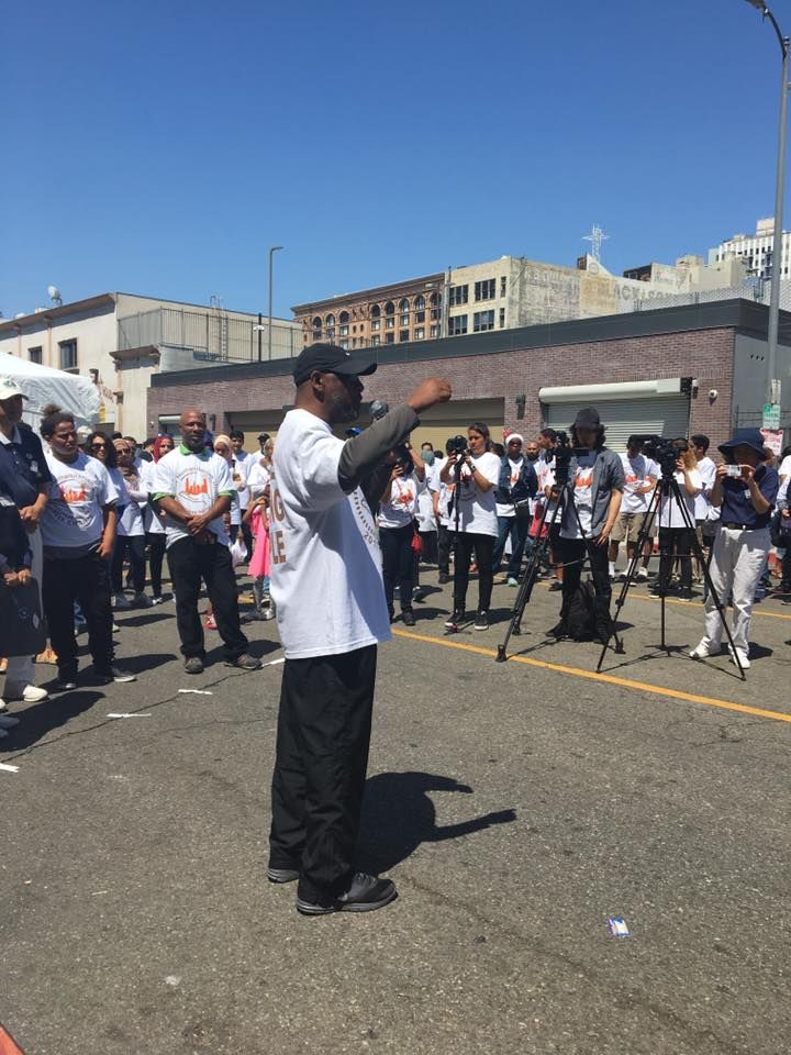 Man speaking to a crowd on a city street; crowd wears white shirts.