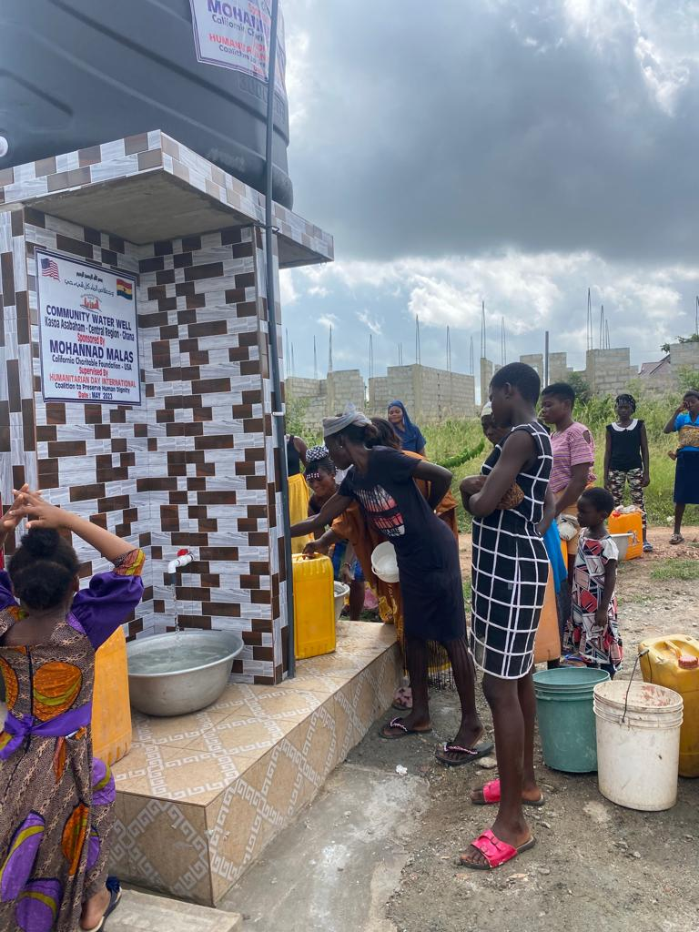 People filling containers at a water tap in front of a water tower, some are waiting.
