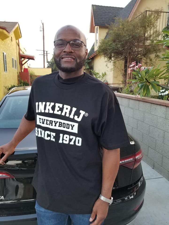 Man in black t-shirt with text stands by a car in front of houses; smiling.