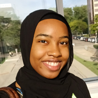 Woman with a black hijab smiling, in front of a window with a city view.