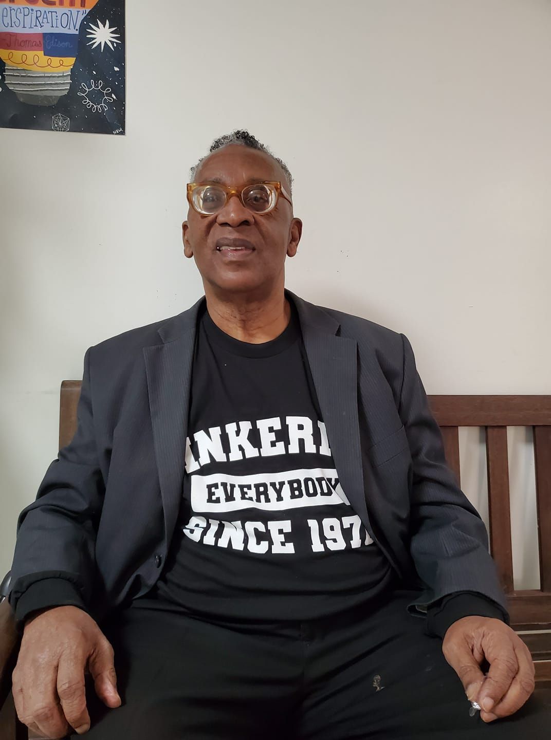 Man wearing glasses and a black t-shirt with a jacket, sitting, smiling.