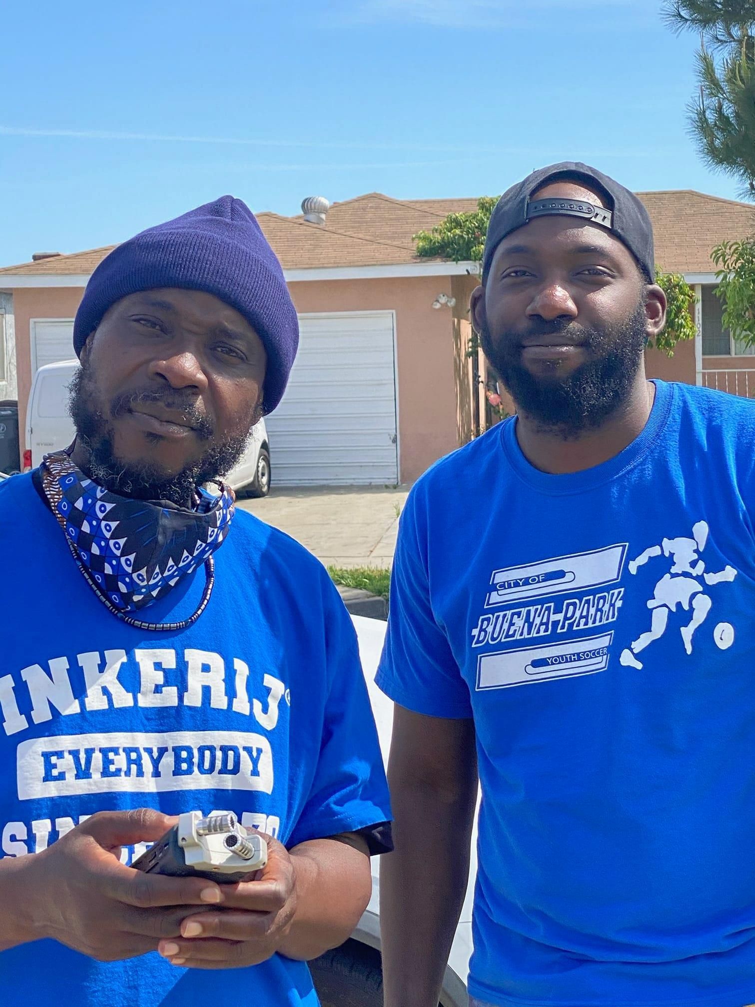 Two men in blue shirts pose outdoors. One holds a small object, the other has a black cap. Bright sunny day.