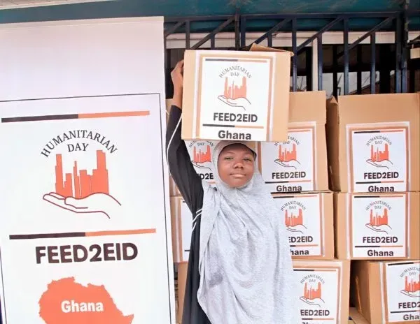 Woman balances food box on her head, standing near boxes labeled 