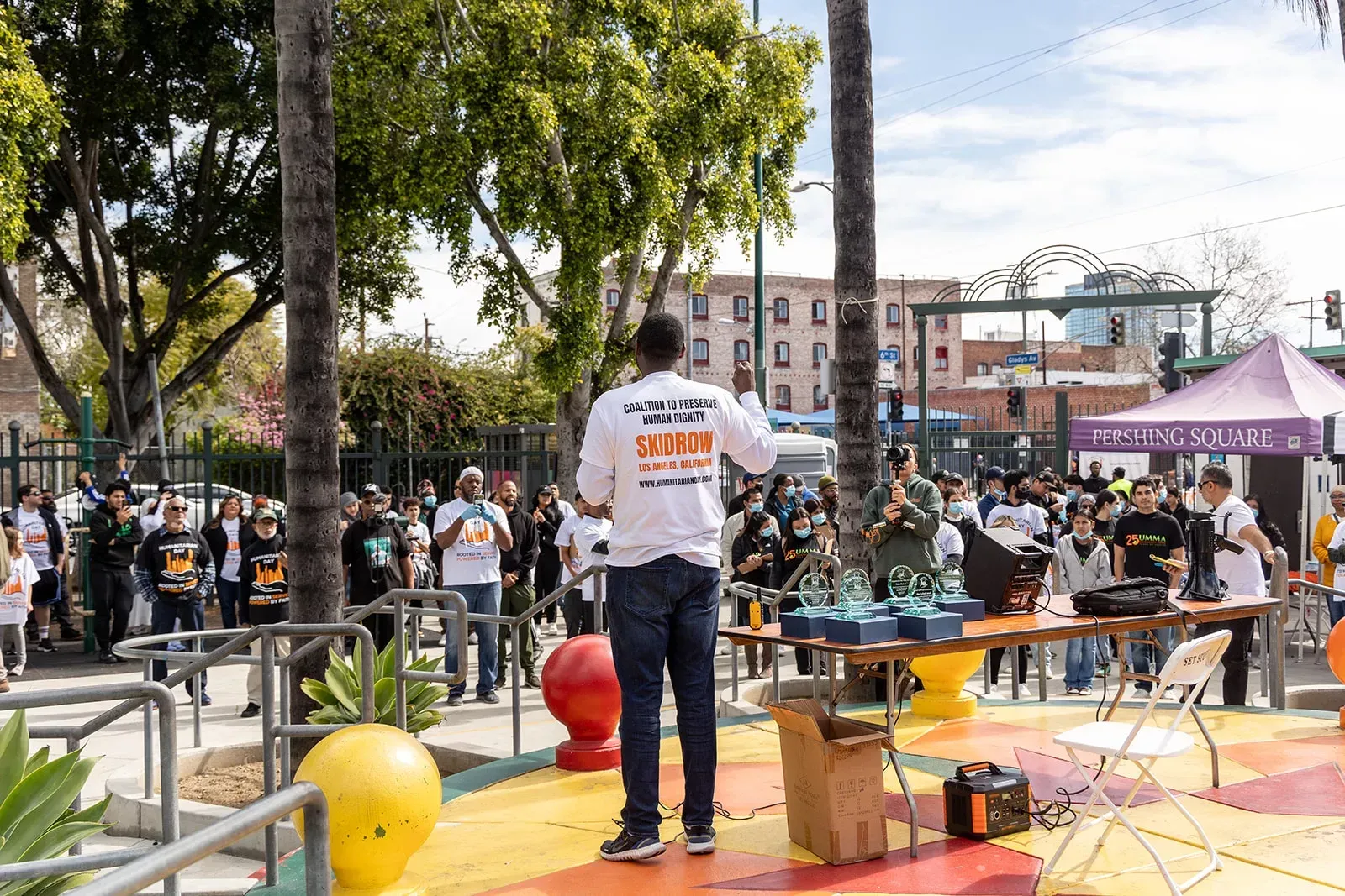 Man addressing a crowd outdoors. People standing near tables with trophies, colorful ground.