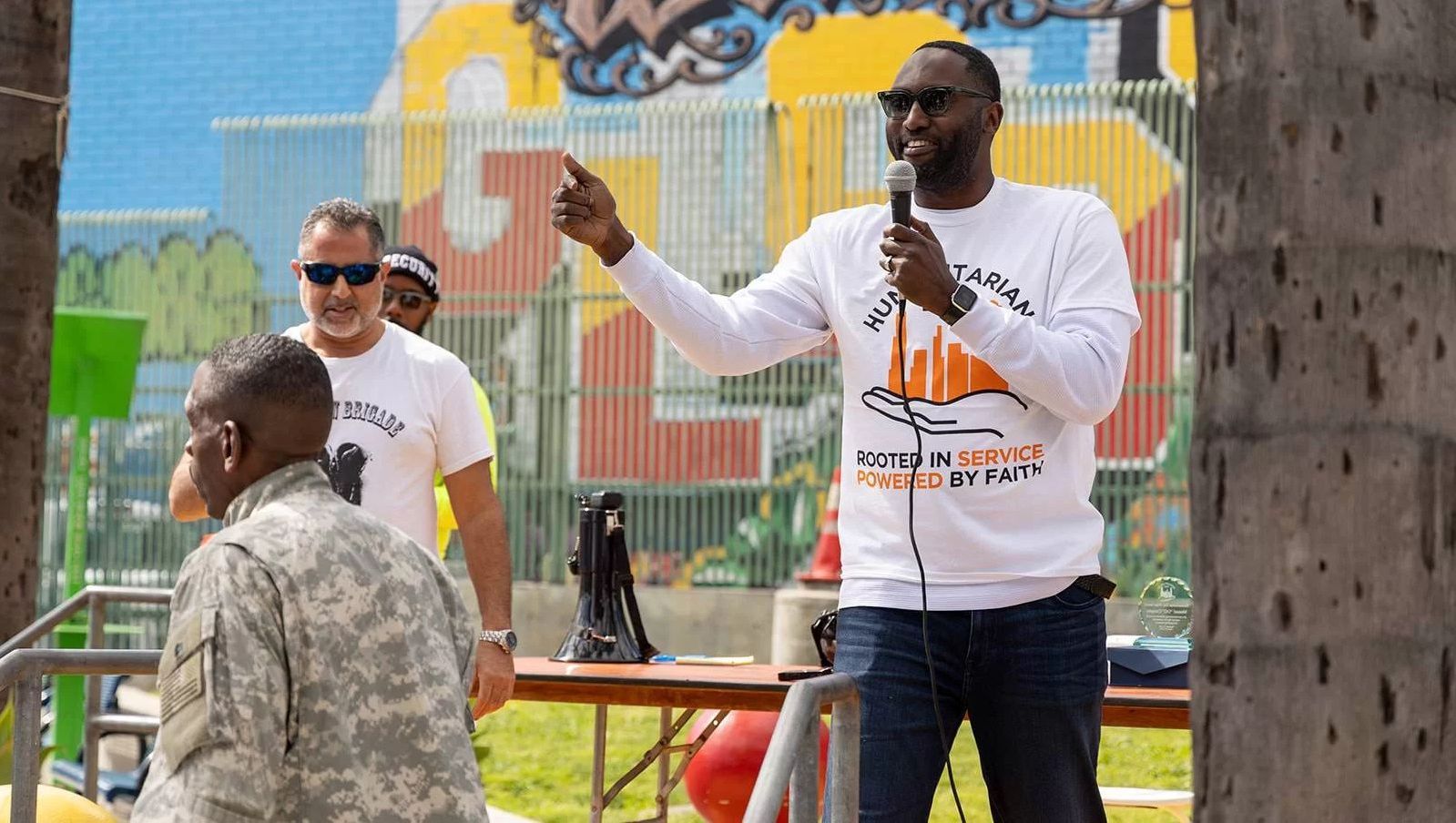 Man speaking into a microphone outdoors, pointing. Others stand nearby. Graffiti art background.