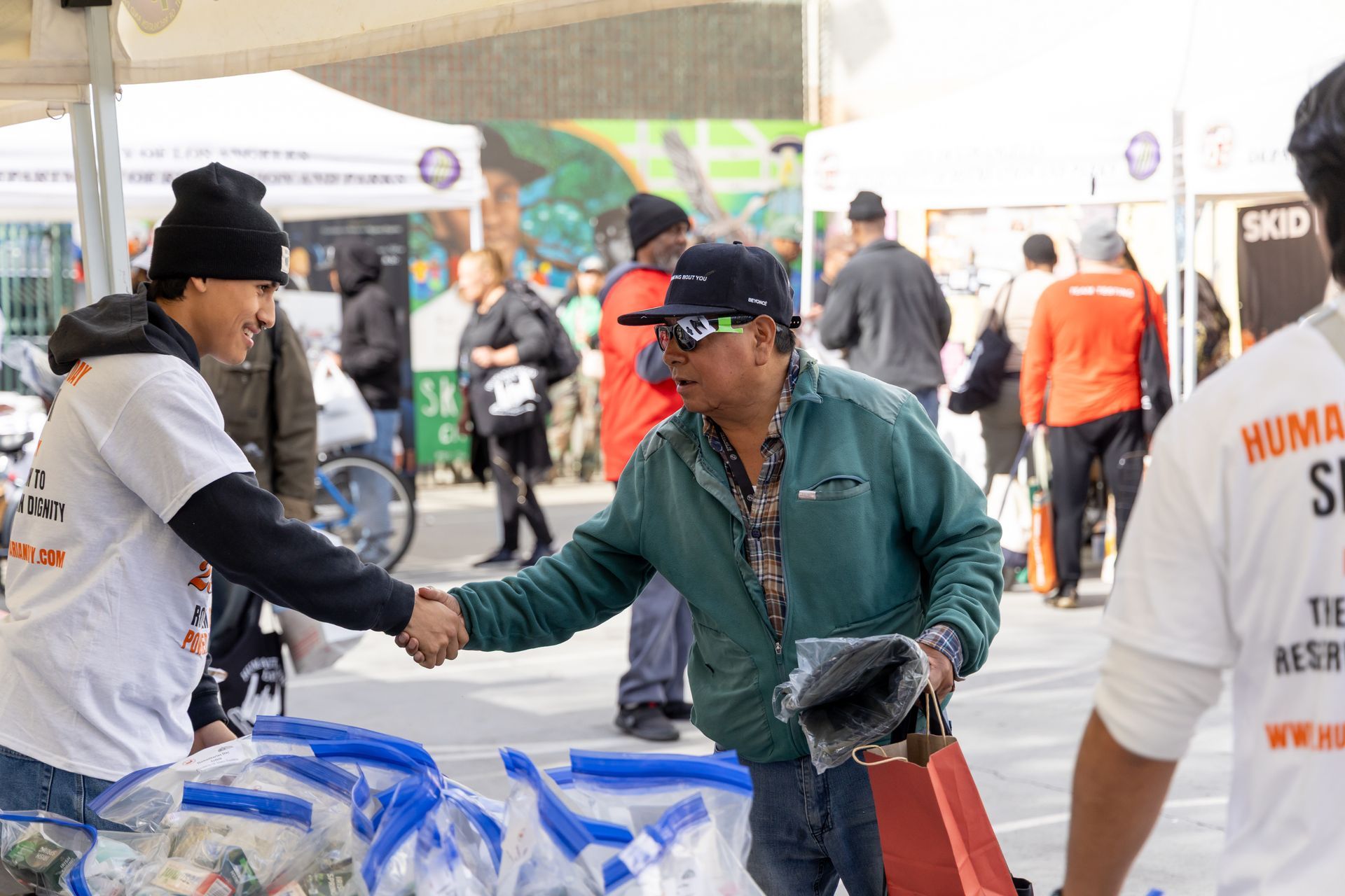 Woman in white shirt shakes hands with man at an outdoor event with tents and bags of supplies.