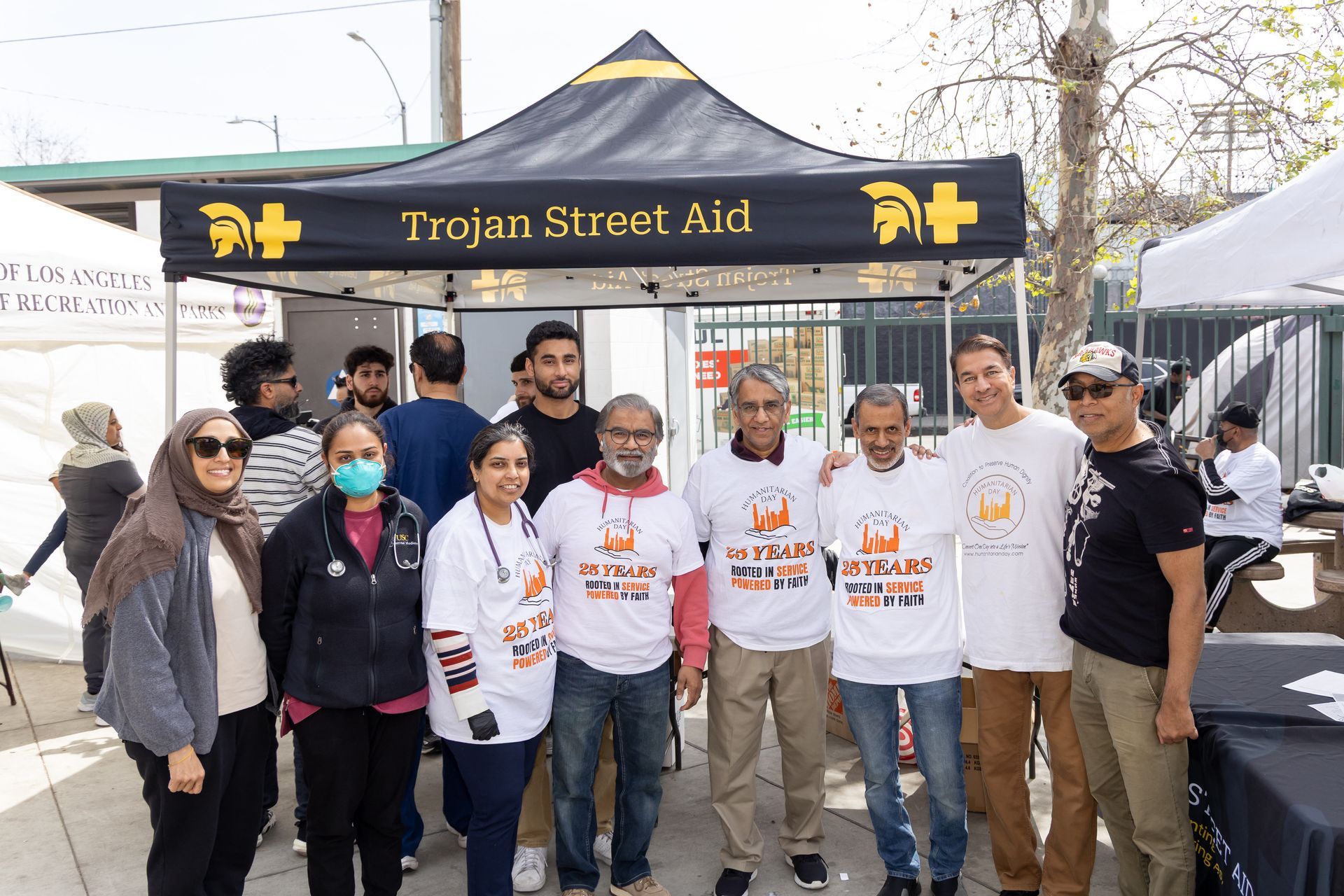Group of people pose in front of a Trojan Street Aid tent, smiling. Some wear t-shirts. Sunny outdoor setting.