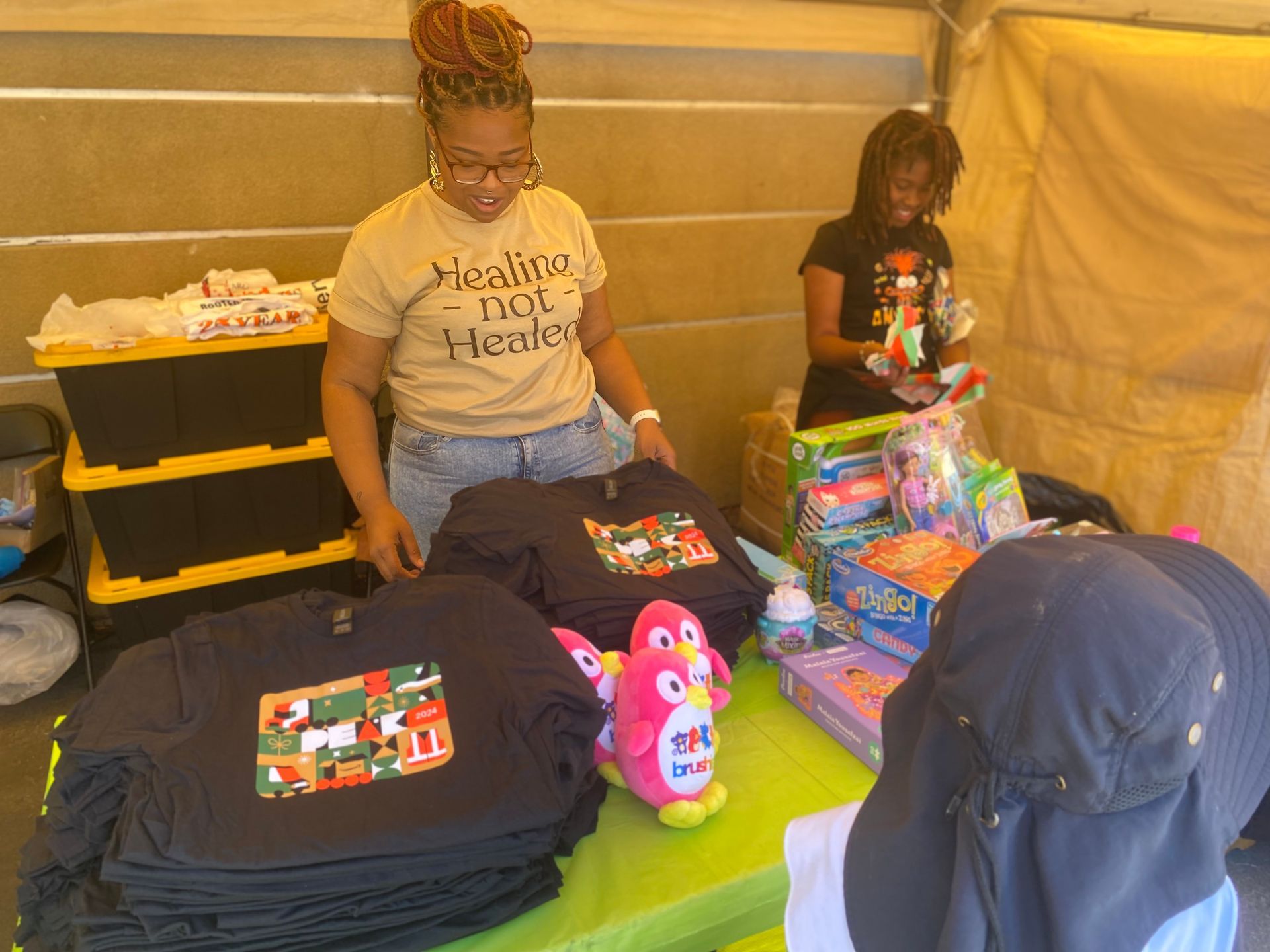 Two women stand behind a vendor table with t-shirts and toys. One woman smiles and looks at the items.