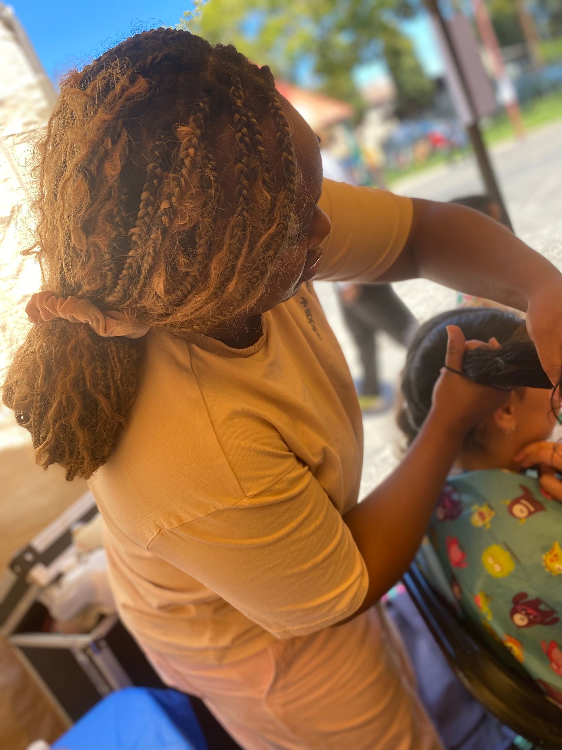 Person in tan shirt cutting someone's hair outdoors; sunny.
