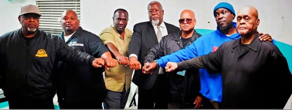 A group of eight Black men standing close together, fists touching, in front of a turquoise wall.