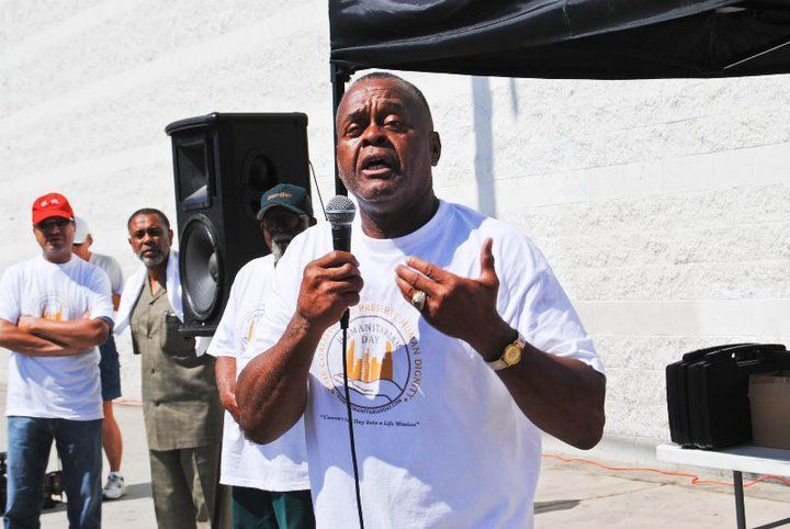 Man speaking into a microphone, gesturing, wearing a white t-shirt, standing outdoors with a crowd.