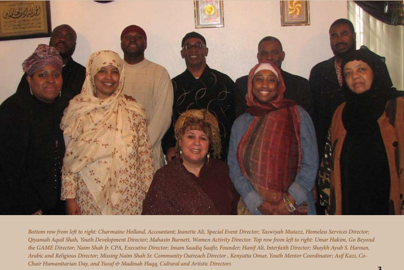 Group of people posing for a photo indoors. People smiling, wearing diverse attire. Artwork on wall behind them.