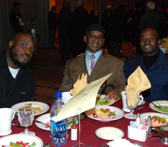 Three men at a table with plates of food, smiling. Well-dressed in a formal setting, possibly a restaurant.