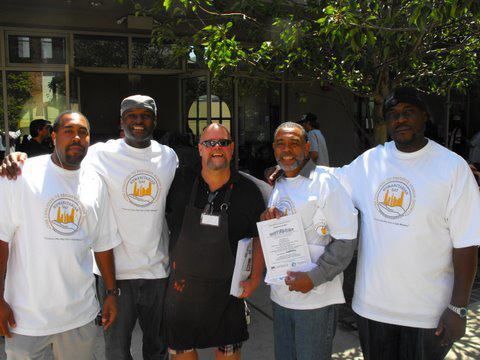 Five men in white shirts, standing outside, smiling. One man holds a document.