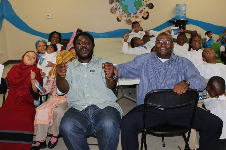 Two men seated in chairs smiling, surrounded by children raising arms in a decorated room.