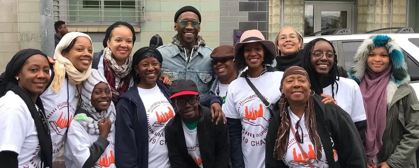 A group of diverse people smile and pose outdoors, some wearing matching white t-shirts with a design.