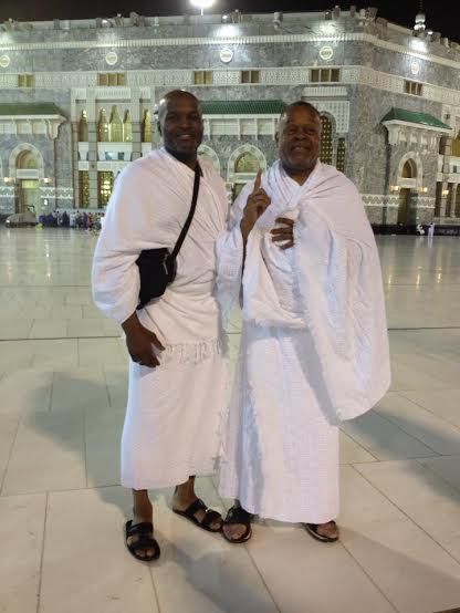 Two men in white robes at the Grand Mosque in Mecca. One points up; both smile.