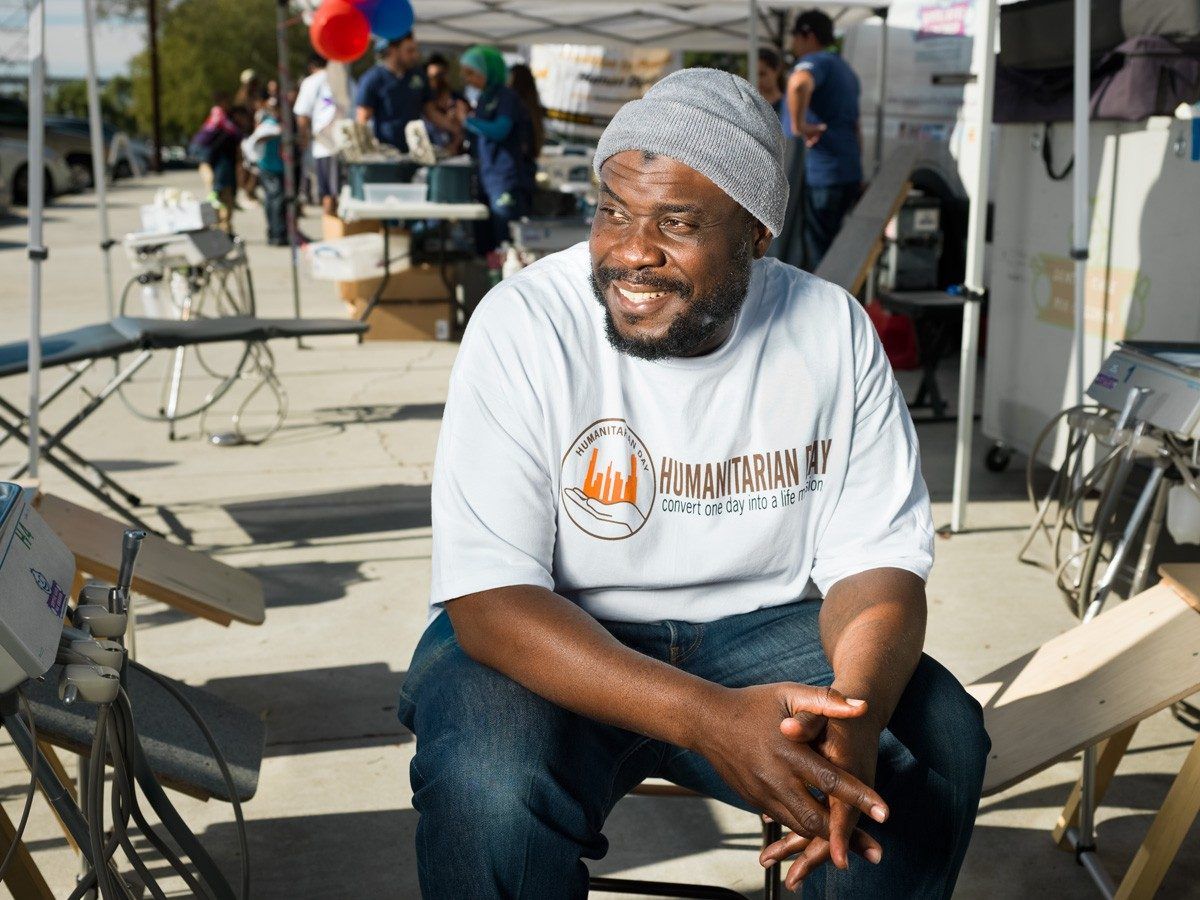 Man wearing a gray hat and white shirt sits outdoors, smiling. Background includes tents and people.