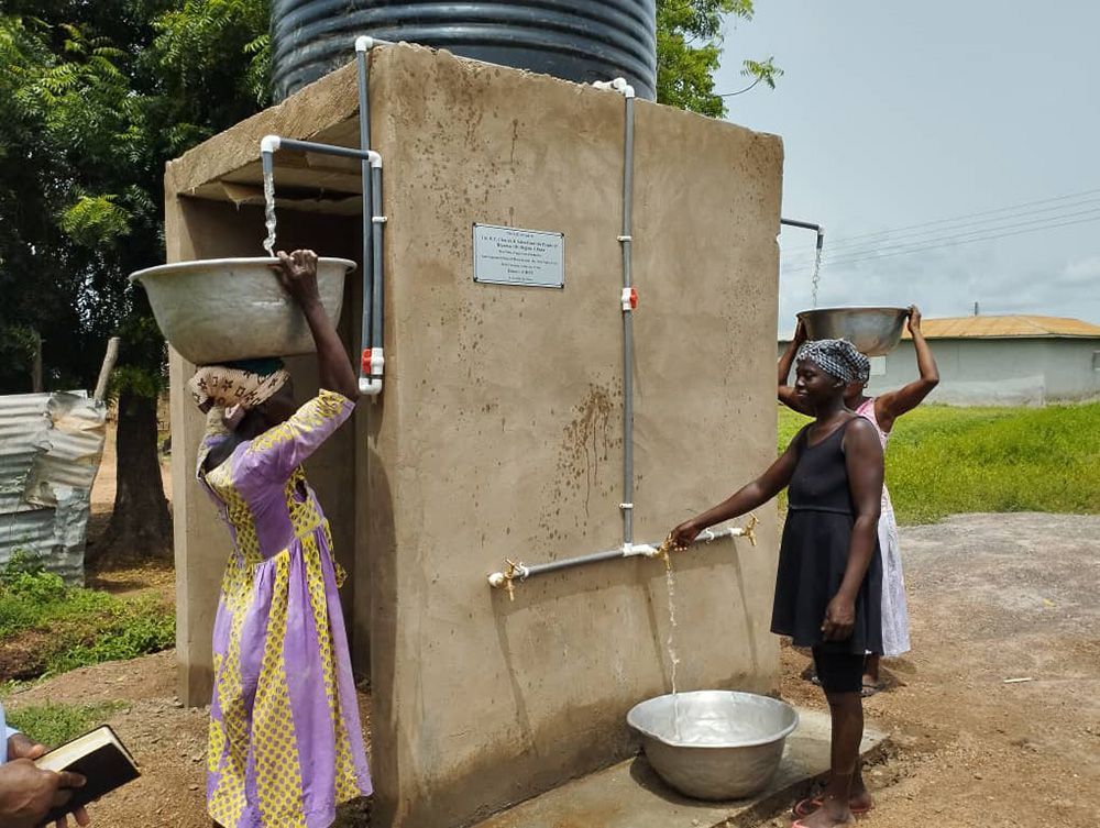 Women fill bowls with water from a tap near a tank. Light concrete structure, outdoors, sunny.
