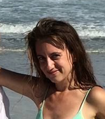 Woman with brown hair smiles in front of the ocean; wearing a light green tank top.