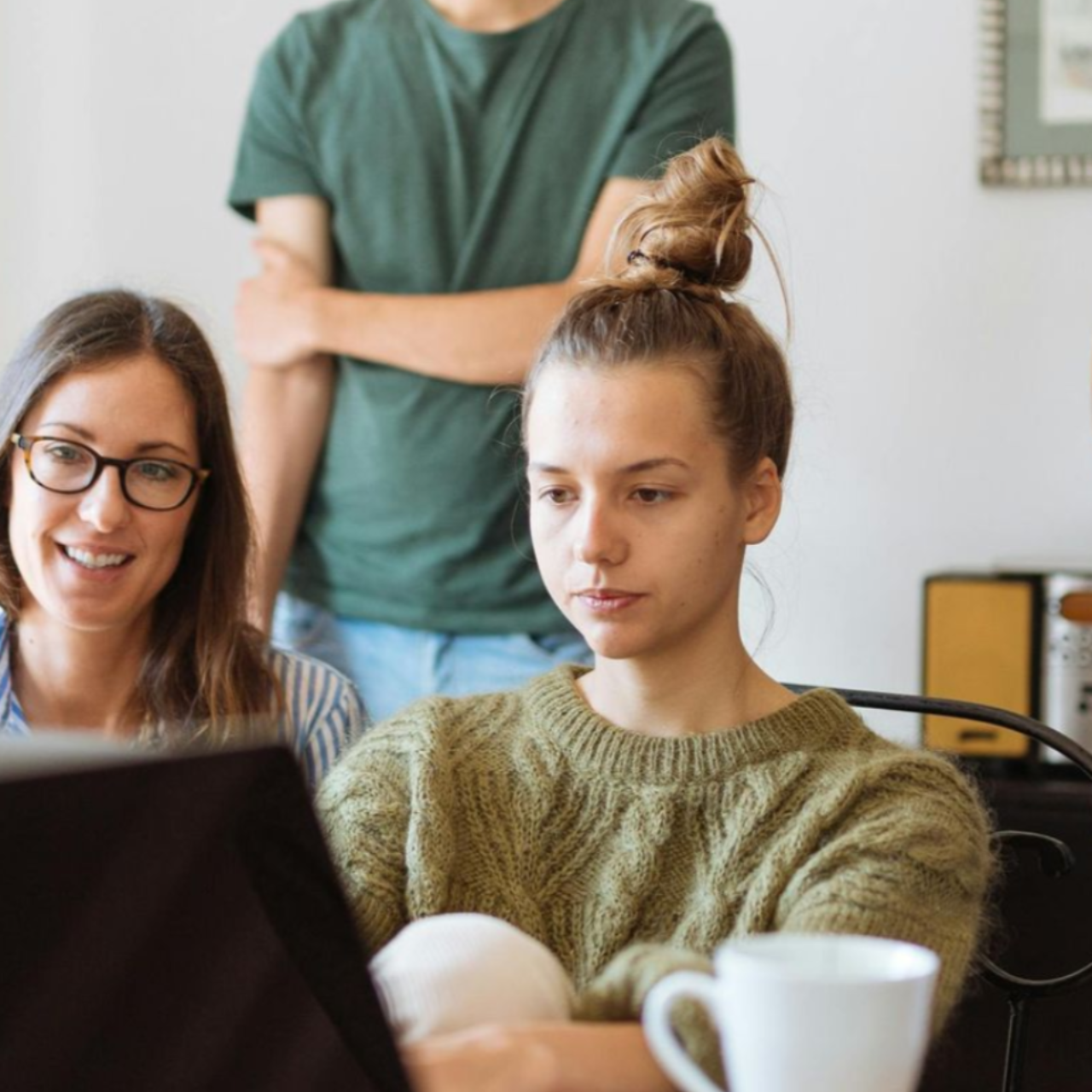 Three people looking at laptop, one woman wears glasses, another a green sweater, third stands behind.