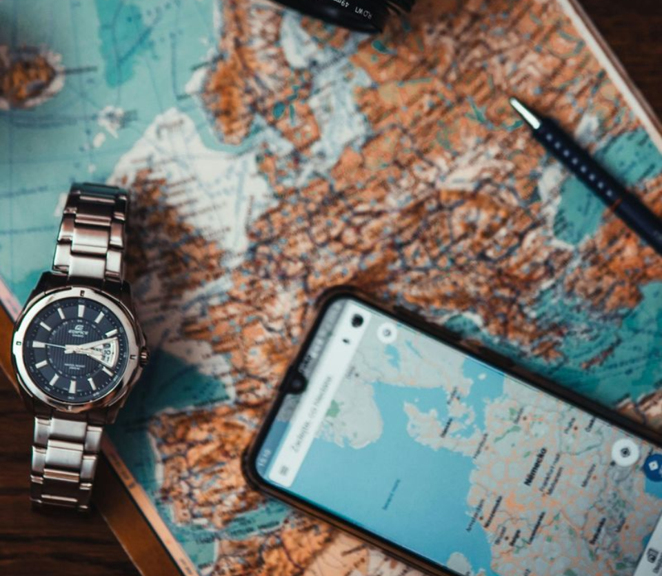 Watch, phone with map, pen, and map of Europe on a wood surface.