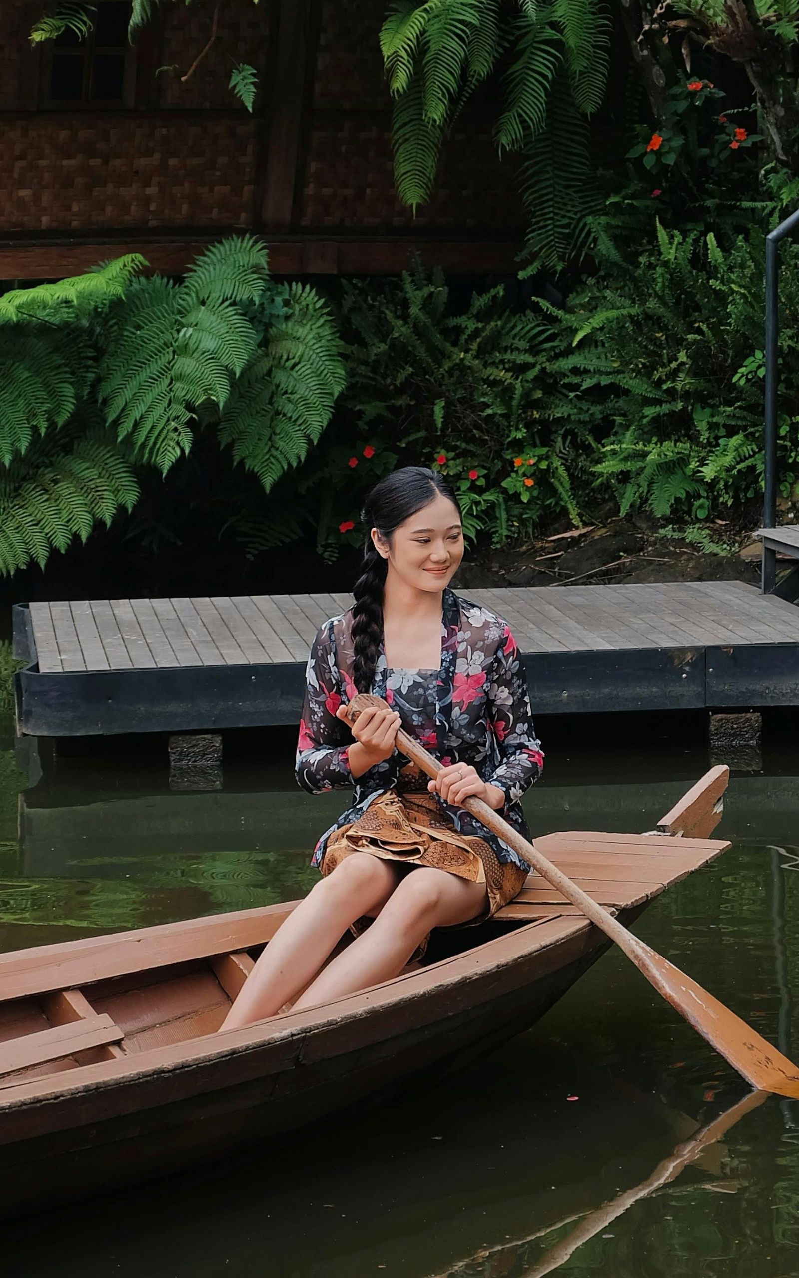 Woman rowing a wooden boat on a river, smiling. Lush green foliage and wooden structure in the background.