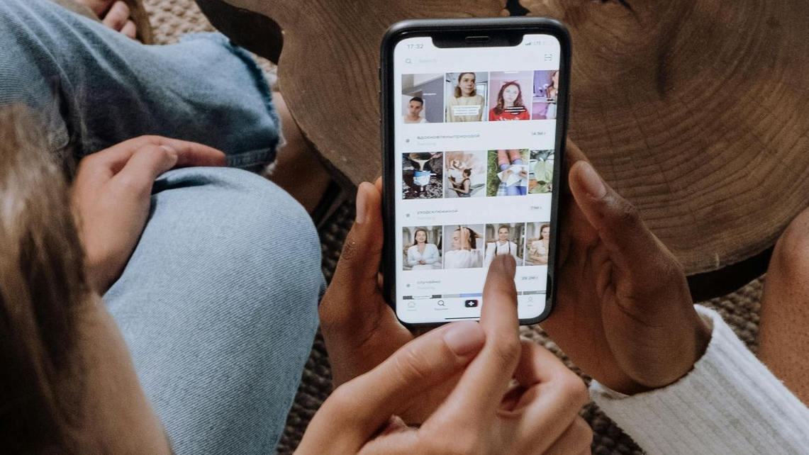 Hands holding a phone, scrolling through images. A person in jeans and a white sweater is also visible.