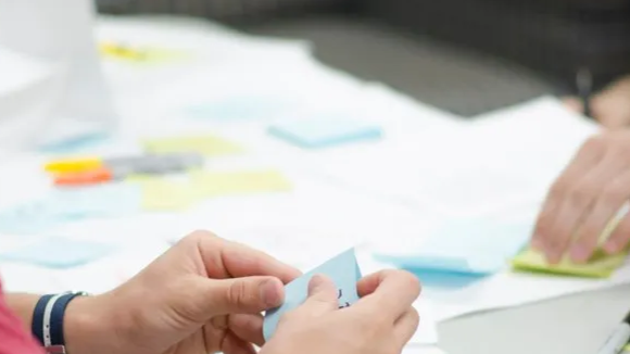 Hands holding a blue sticky note at a white table with other notes; blurred background.