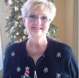 Woman with short white hair, glasses, smiles, wearing a black sweater with snowflakes, in front of a Christmas tree.