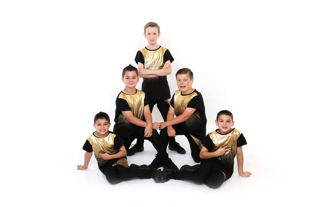 A dance group of five boys in gold and black outfits poses on a white background.