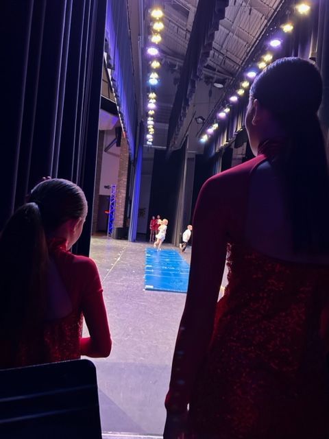 Two girls in red dresses watch a performance from the wings of a theater.