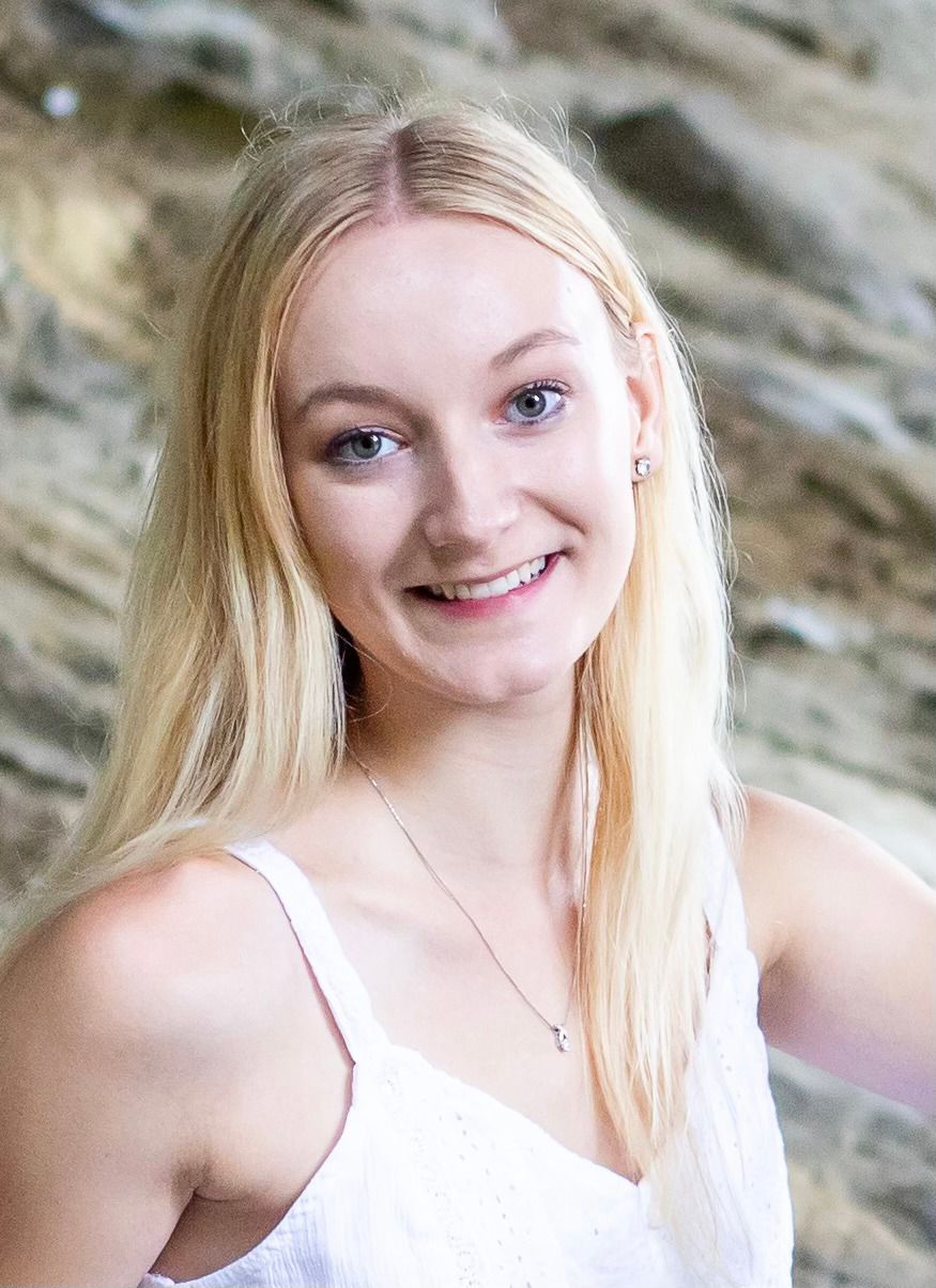Blonde woman smiling, wearing a white top, posing in front of a textured, light-colored background.