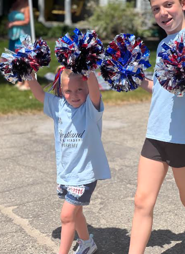 Two children with pom-poms, a young girl cheering in light blue shirt and shorts, outdoors.