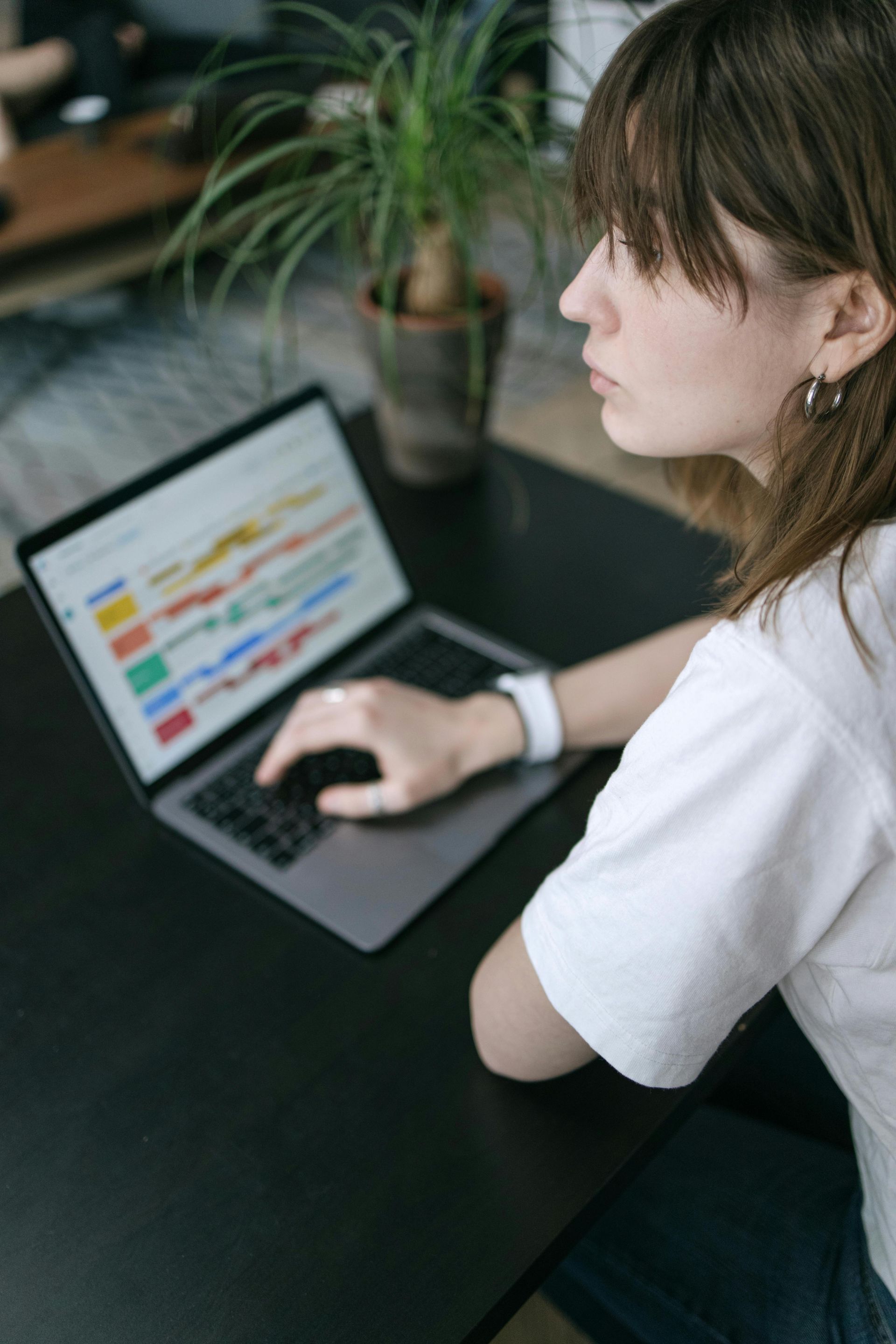 A person in a white shirt sits at a dark table, focused on a laptop screen displaying colorful data charts.