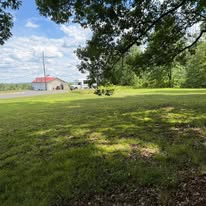 Grassy field with trees and a small building under a partly cloudy sky