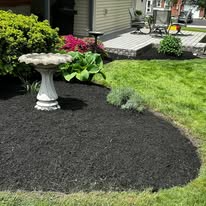 Mulched flower bed with birdbath, green lawn, and house porch in the background
