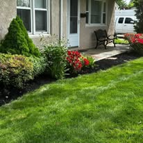 Front yard garden with red flowers, green lawn, and a porch bench beside a house