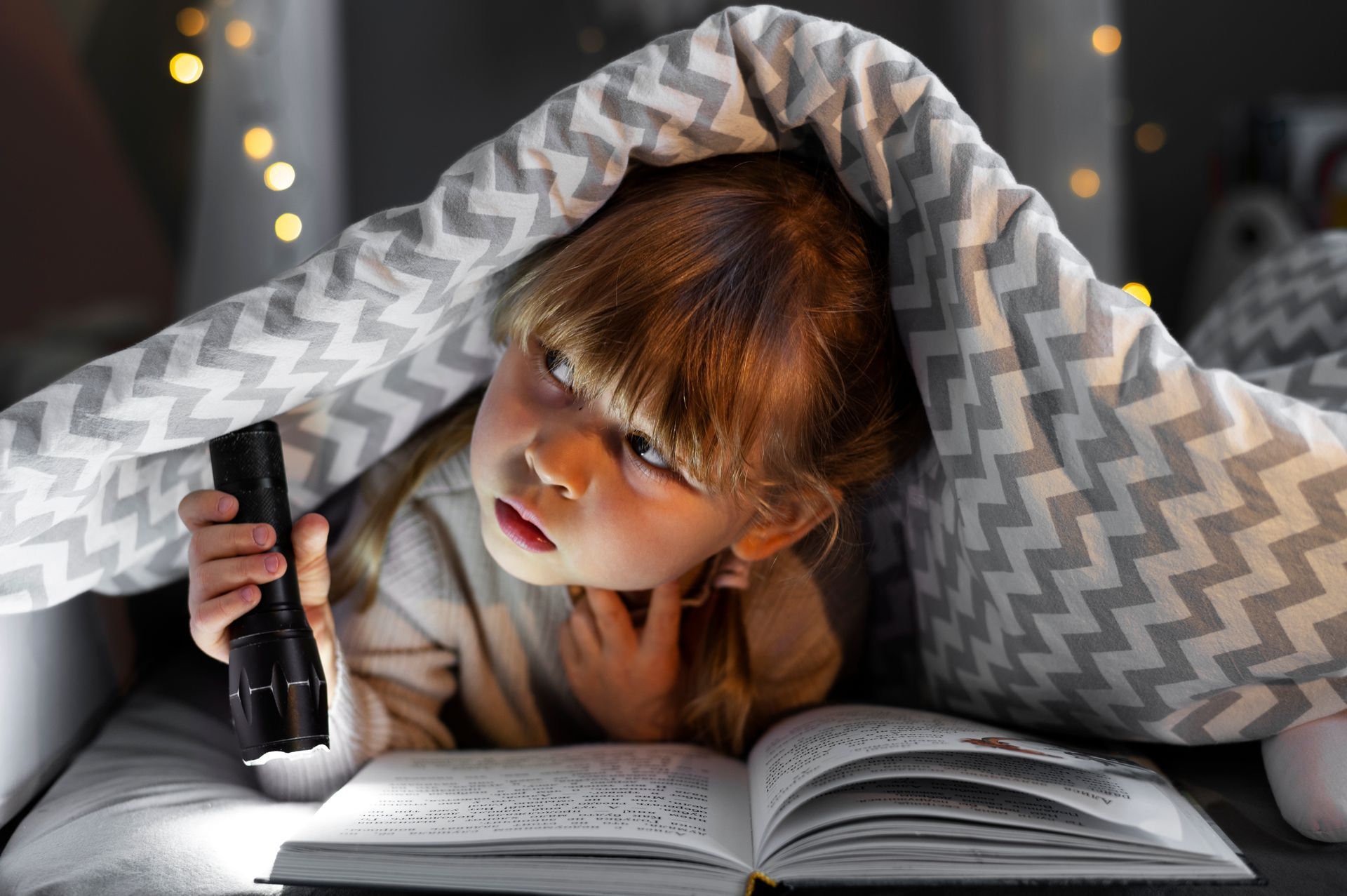 Jeune enfant lisant sous une couverture, utilisant une lampe de poche, expression concentrée.