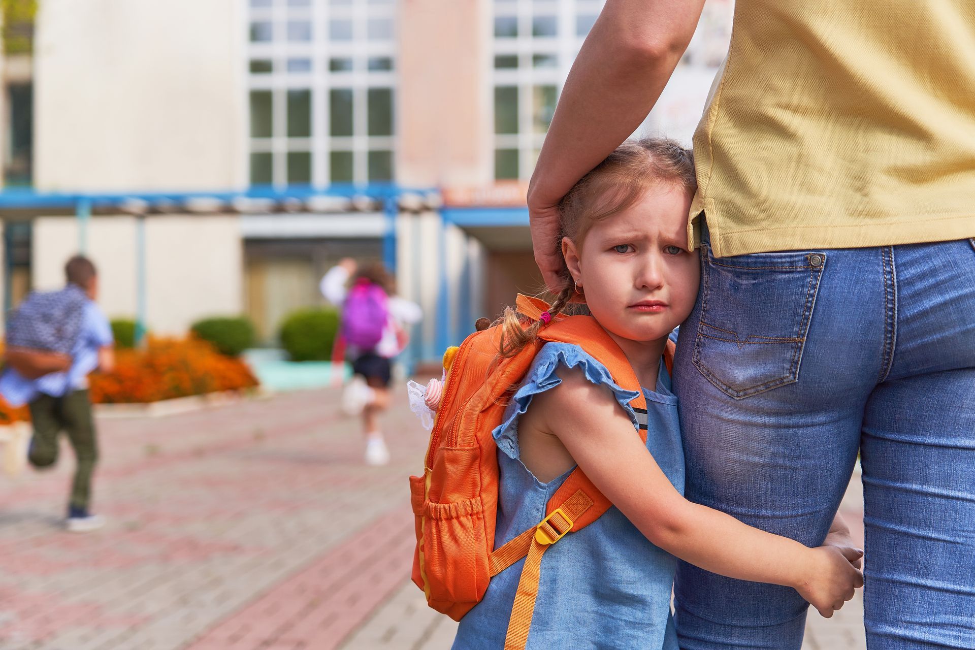 Une enfant s'accroche à la jambe d'un adulte, l'air triste, près d'un bâtiment scolaire, tandis que d'autres enfants courent.