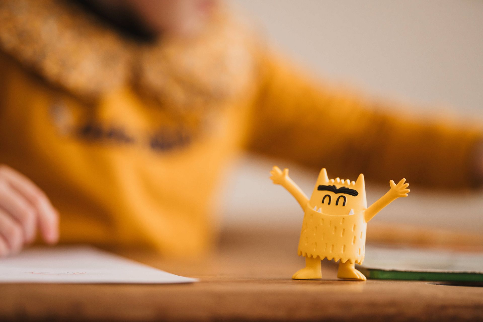 Une figurine de monstre jaune, les bras levés, posée sur une table en bois, devant un enfant vêtu de jaune.